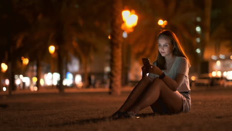 A-beautiful-young-girl-in-the-summer-in-a-big-night-city-keeps-a-smart-phone-in-shell-and-writes-a-message-reads-e-mail-and-communicates-in-chat-rooms-and-social-networks-keeps-a-blog-looks-at-the-phone-screen